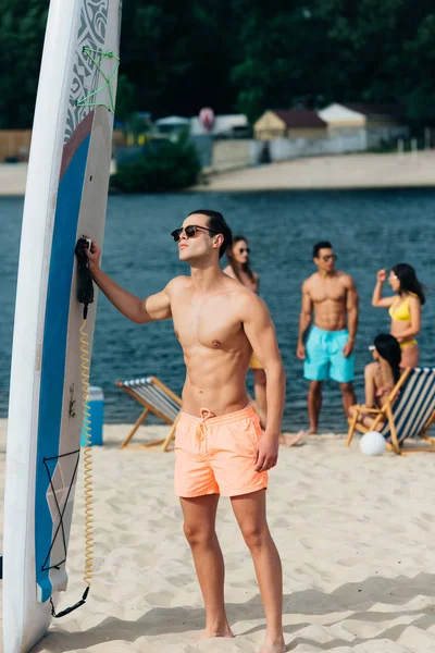 Schöner junger Mann mit Sonnenbrille steht neben Surfbrett am Strand — Stockfoto