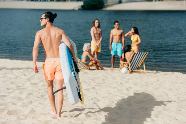 Vista trasera del joven sosteniendo tabla de surf mientras camina en la playa cerca de amigos multiculturales - foto de stock