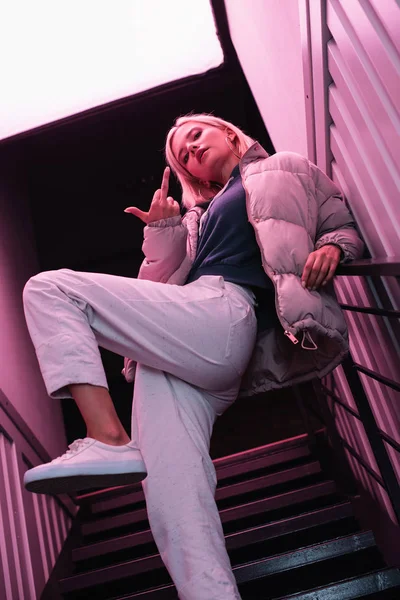 Vista inferior de la chica rubia con estilo en la chaqueta de pie en las escaleras y mostrando el dedo medio - foto de stock