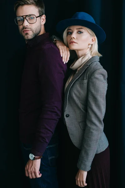 Stylish young man in glasses and blonde woman in hat looking at camera near curtain — Stock Photo