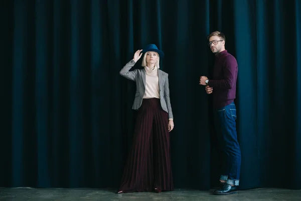 Vista completa de hombre joven con estilo en gafas y mujer rubia en sombrero cerca de la cortina - foto de stock