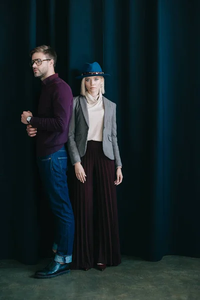 Vista completa de hombre joven con estilo en gafas y mujer rubia en sombrero cerca de la cortina - foto de stock