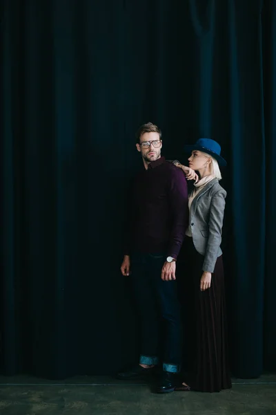 Full length view of stylish young man in glasses and blonde woman in hat near curtain — Stock Photo
