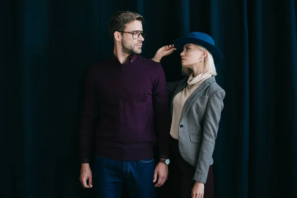 Joven con estilo en gafas y mujer rubia en sombrero mirándose cerca de la cortina - foto de stock