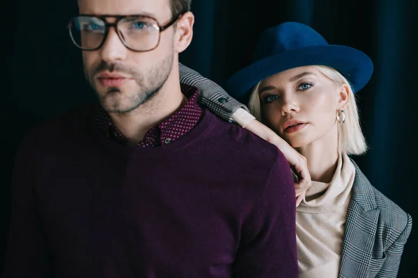 Joven con estilo en gafas y mujer rubia en sombrero mirando a la cámara cerca de la cortina - foto de stock