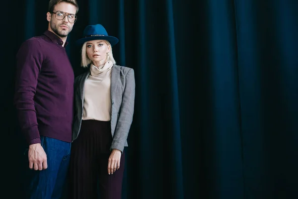 Élégant jeune homme dans des lunettes et femme blonde dans le chapeau regardant la caméra près du rideau — Photo de stock