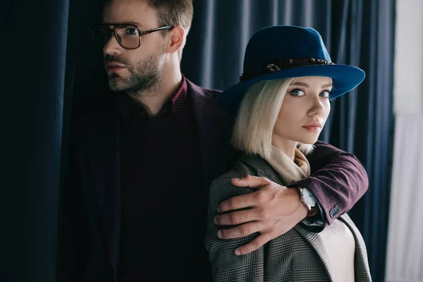 Elegante joven en gafas abrazando chica rubia en sombrero cerca de la cortina - foto de stock