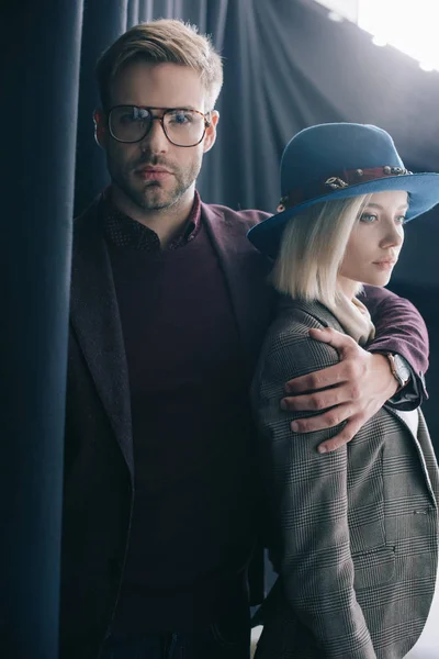 Elegant young man in glasses embracing blonde girl in hat near curtain — Stock Photo