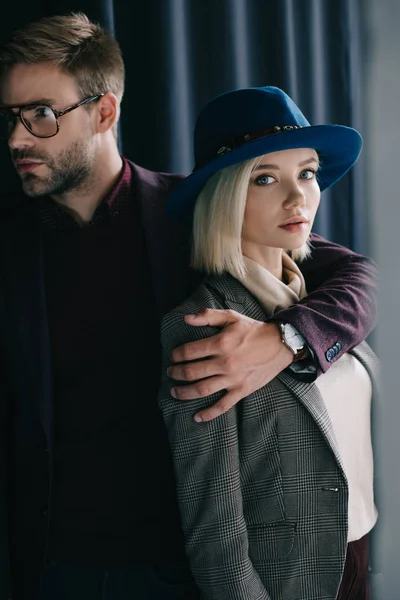 Elegant young man in glasses embracing blonde girl in hat near curtain — Stock Photo