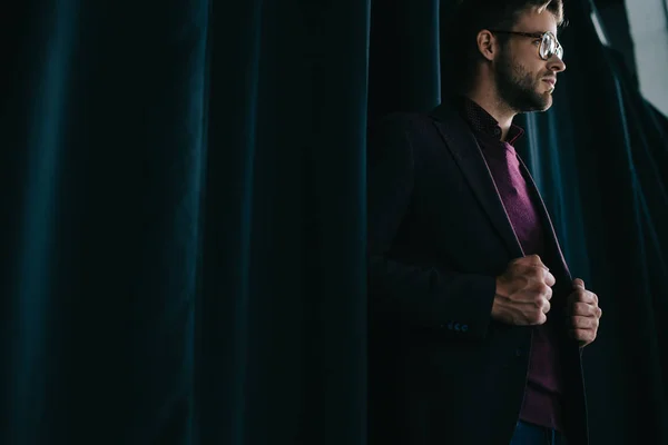 Stylish young man in jacket and glasses standing near curtain and looking away — Stock Photo