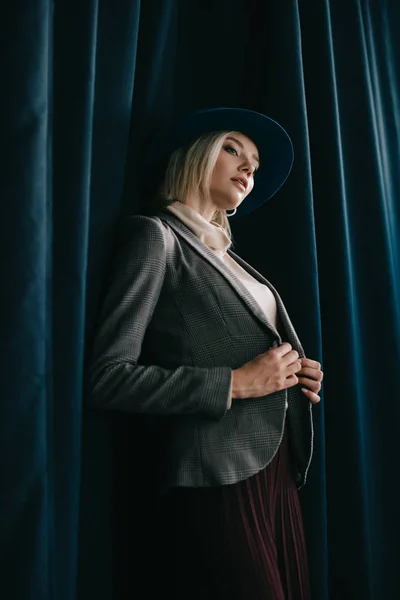 Стильная блондинка в шляпе и куртке, стоящая возле занавеса — стоковое фото