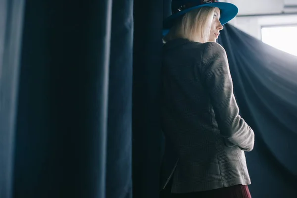 Mujer rubia elegante en sombrero y chaqueta de pie cerca de la cortina - foto de stock