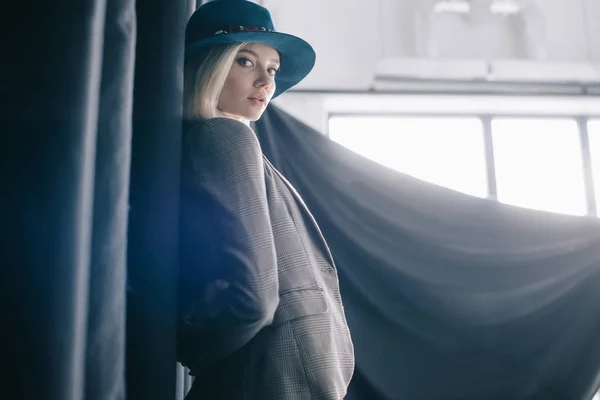 Elegante chica rubia en sombrero y chaqueta de pie cerca de la cortina y mirando a la cámara - foto de stock