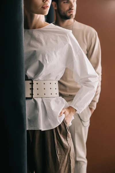 Обрізаний вид на стильну молоду жінку в блузці і чоловіка, що стоїть біля завіси на коричневому — стокове фото