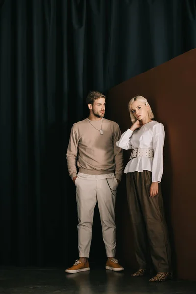 Visão comprimento total do homem elegante com as mãos em bolsos e mulher loira em blusa de pé perto da cortina — Fotografia de Stock
