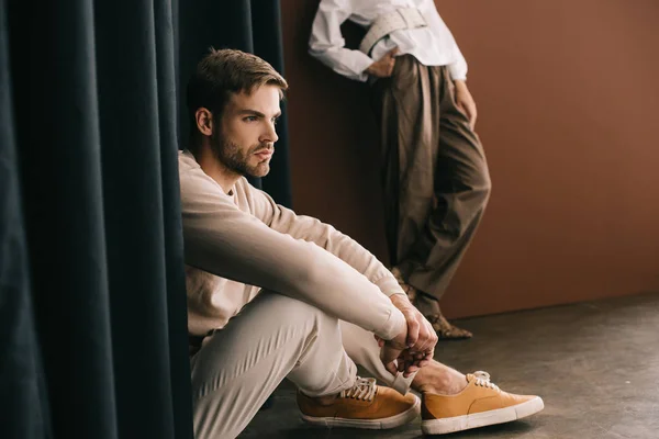 Ausgeschnittene Ansicht einer Frau, die mit der Hand auf der Hüfte steht, und eines bärtigen Mannes, der neben einem Vorhang sitzt — Stockfoto