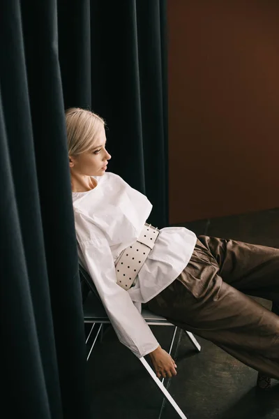 Stylische blonde Frau in weißer Bluse sitzt auf Stuhl neben Vorhang auf braun — Stockfoto