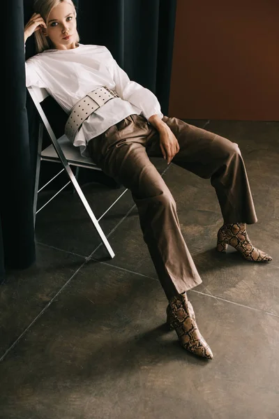 Стильна блондинка в білій блузці і чоботи зі змією, що сидить на стільці біля завіси на коричневому — Stock Photo