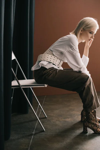 Vista laterale di elegante donna bionda in camicetta bianca e stivali con stampa di pelle di serpente seduta sulla sedia vicino alla tenda sul marrone — Foto stock