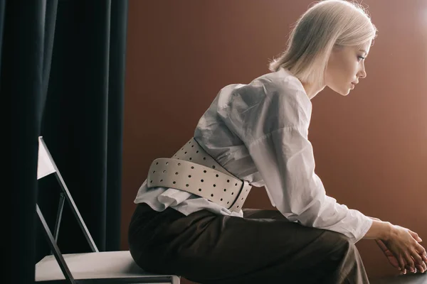 Вид сбоку стильной блондинки в белой блузке, сидящей на стуле возле занавески на коричневом — стоковое фото