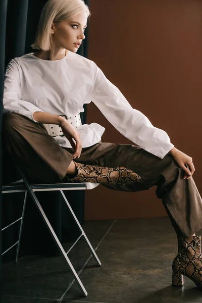 Mulher loira elegante em blusa branca e botas com impressão de pele de cobra sentado na cadeira perto de cortina em marrom — Fotografia de Stock