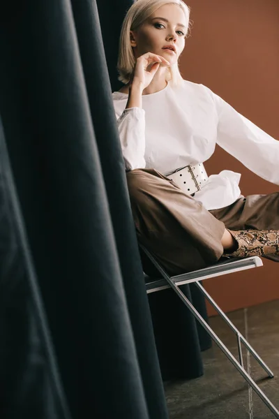 Mujer rubia con estilo en blusa blanca sentada en la silla cerca de la cortina en marrón - foto de stock