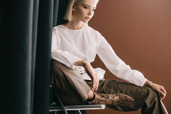 Mulher loira elegante em blusa branca sentado na cadeira perto de cortina em marrom — Fotografia de Stock