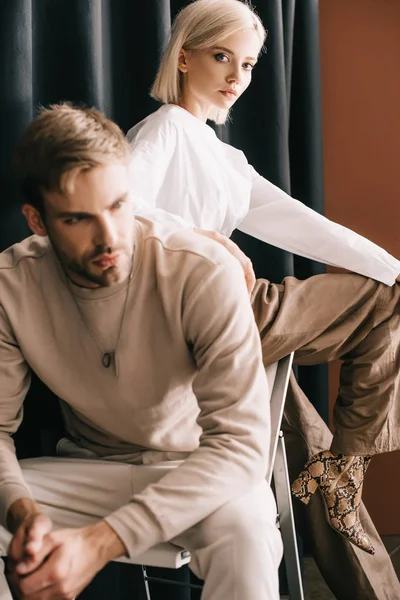 Femme blonde en chemisier blanc et homme barbu assis sur une chaise près du rideau — Photo de stock