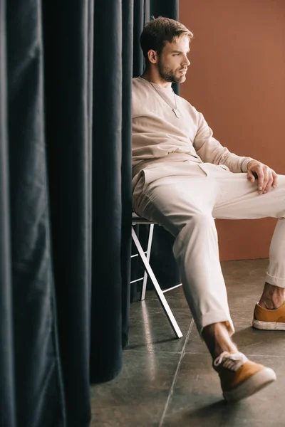Uomo barbuto pensieroso in abito casual seduto sulla sedia vicino alla tenda — Foto stock