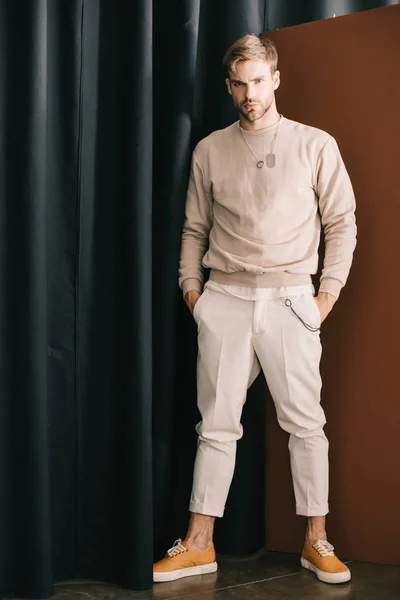 Full length view of serious stylish man standing with hands in pockets near curtain — Stock Photo