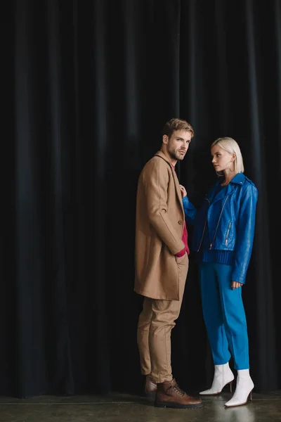 Vista completa de la mujer joven y el hombre con estilo en trajes de otoño cerca de la cortina negra - foto de stock