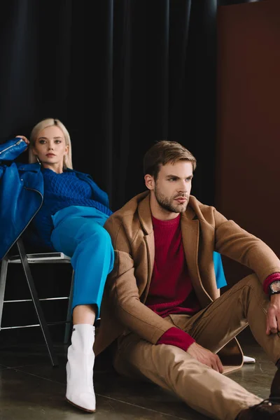 Stylish blonde girl in blue leather jacket sitting on chair near man in brown coat — Stock Photo