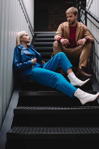 Stylish young woman and man in autumn clothes sitting on stairs and looking at each other — Stock Photo