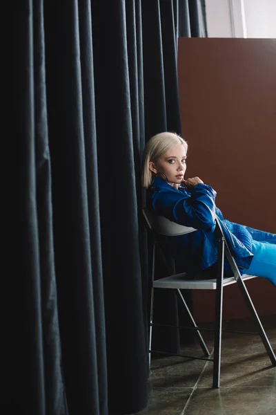 Привлекательная блондинка в кожаной куртке и на каблуках сидит на стуле и смотрит в камеру — стоковое фото