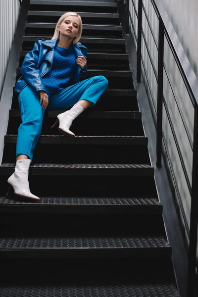 Блондинка в синій шкіряній куртці і на підборах сидить на сходах — стокове фото