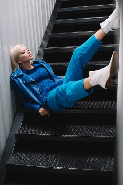 Blonde woman wearing blue leather jacket and heels sitting on stairs — Stock Photo