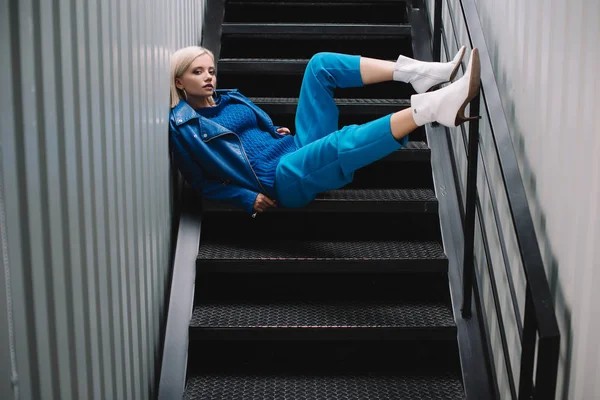 Blonde woman wearing blue leather jacket and heels sitting on stairs — Stock Photo