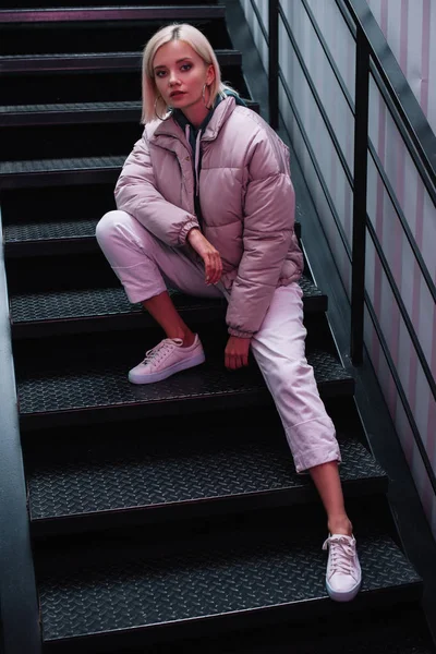 Fille blonde en duvet et baskets assis sur les escaliers et regardant la caméra — Photo de stock