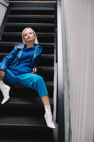 Блондинка в синей кожаной куртке и на каблуках сидит на лестнице и смотрит в камеру — стоковое фото
