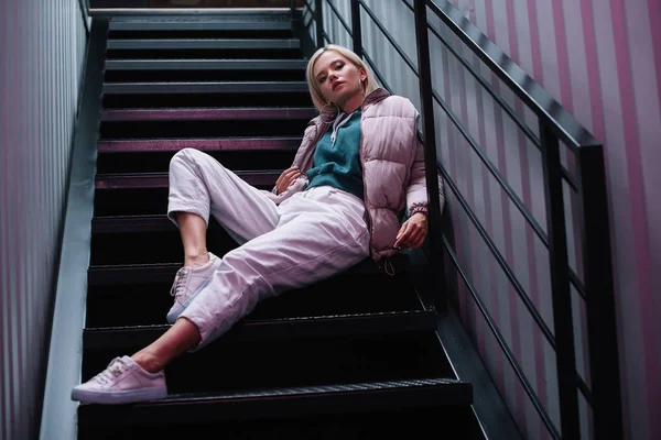 Selbstbewusstes blondes Mädchen in Daunenjacke und Turnschuhen sitzt auf der Treppe — Stockfoto