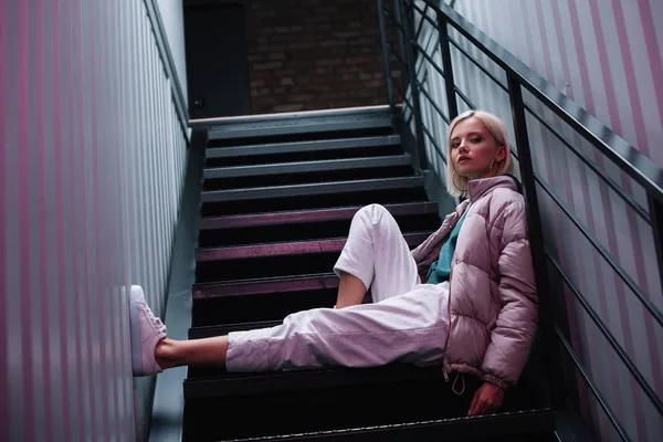 Selbstbewusstes blondes Mädchen in Daunenjacke und Turnschuhen, das auf der Treppe sitzt und in die Kamera blickt — Stockfoto