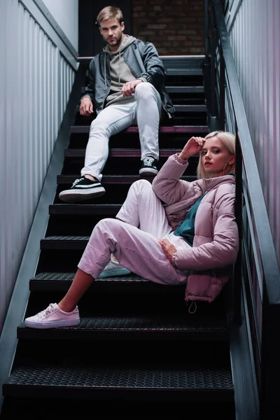 Стильный молодой человек и женщина в повседневных осенних нарядах сидят на лестнице и смотрят в камеру — стоковое фото