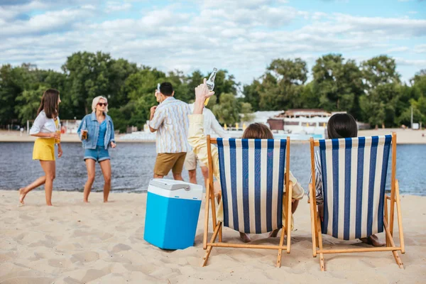 Vue arrière de l'homme et de la femme assis dans des chaises longues près d'amis multiculturels s'amusant sur la plage — Photo de stock