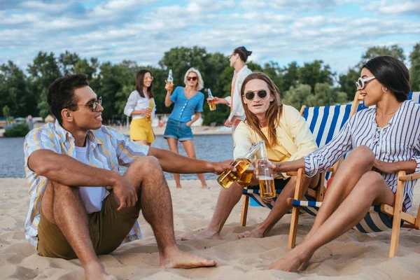 Cheerful multiethnic friends clinking bottles of beer while having fun on beach — Stock Photo