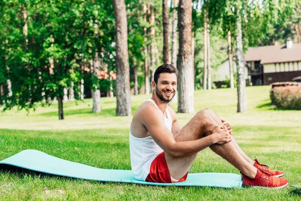 Cheerful bearded sportsman sitting on fitness mat in park — Stock Photo