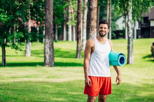 Cheerful bearded man holding fitness mat in park — Stock Photo