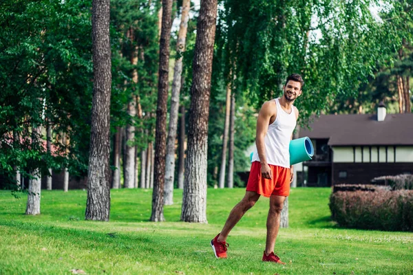 Веселый бородатый мужчина держит фитнес мат во время прогулки в парке — стоковое фото