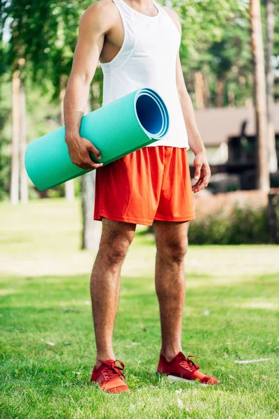 Vista recortada de deportista sosteniendo alfombra de fitness mientras está de pie en la hierba - foto de stock