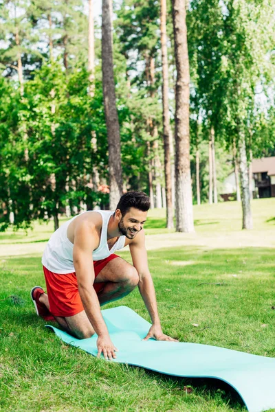 Deportista feliz sonriendo mientras está sentado en la alfombra de fitness en el parque - foto de stock