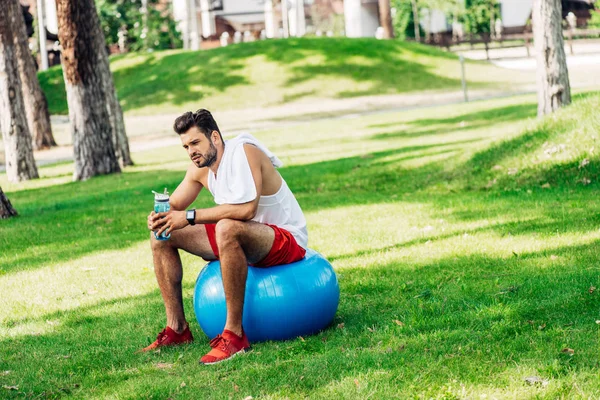 Barbudo deportista sosteniendo botella de deporte mientras está sentado en la pelota de fitness - foto de stock